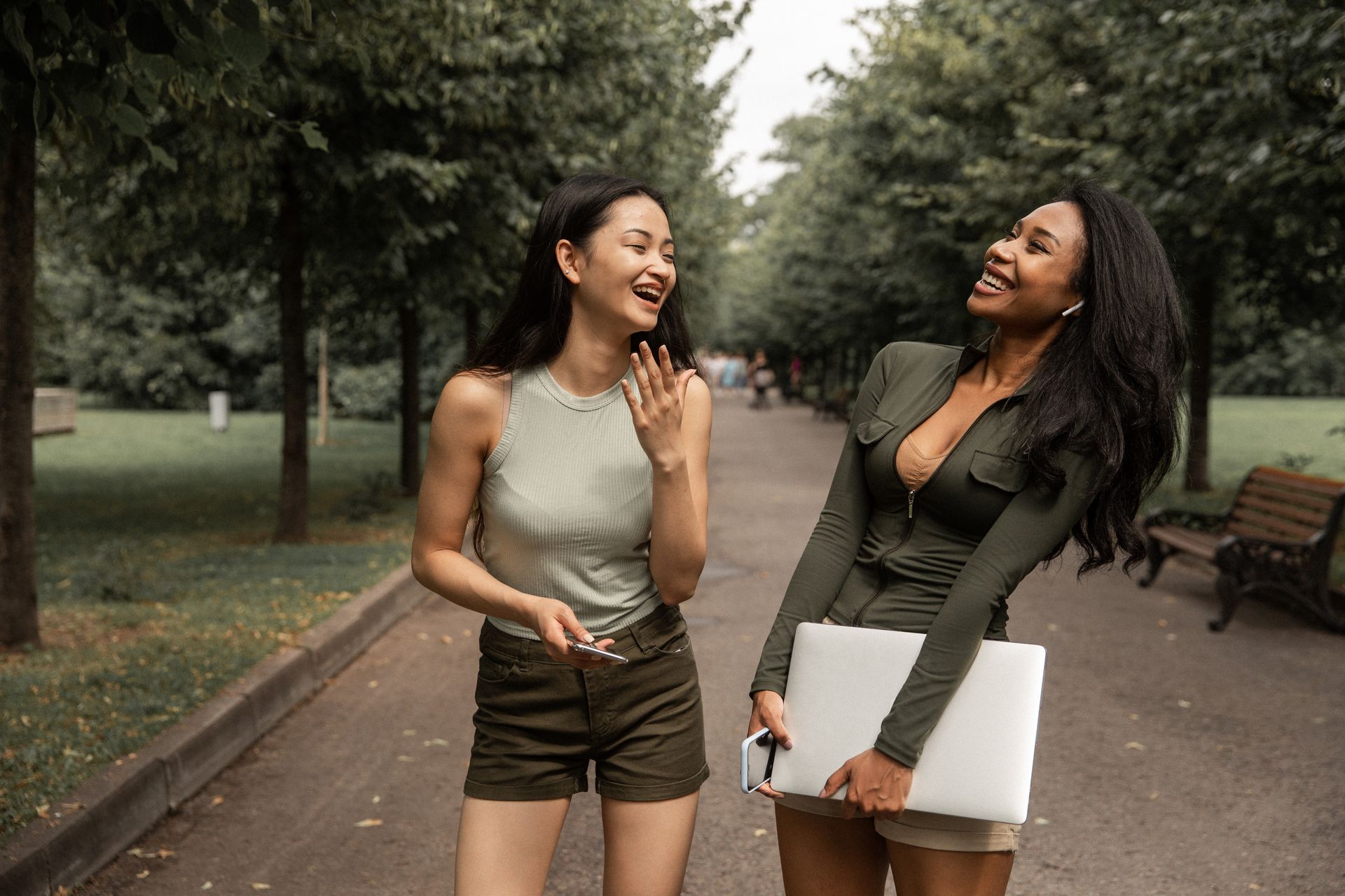 Two young women laugh and enjoy a conversation outdoors in a park on a warm day.