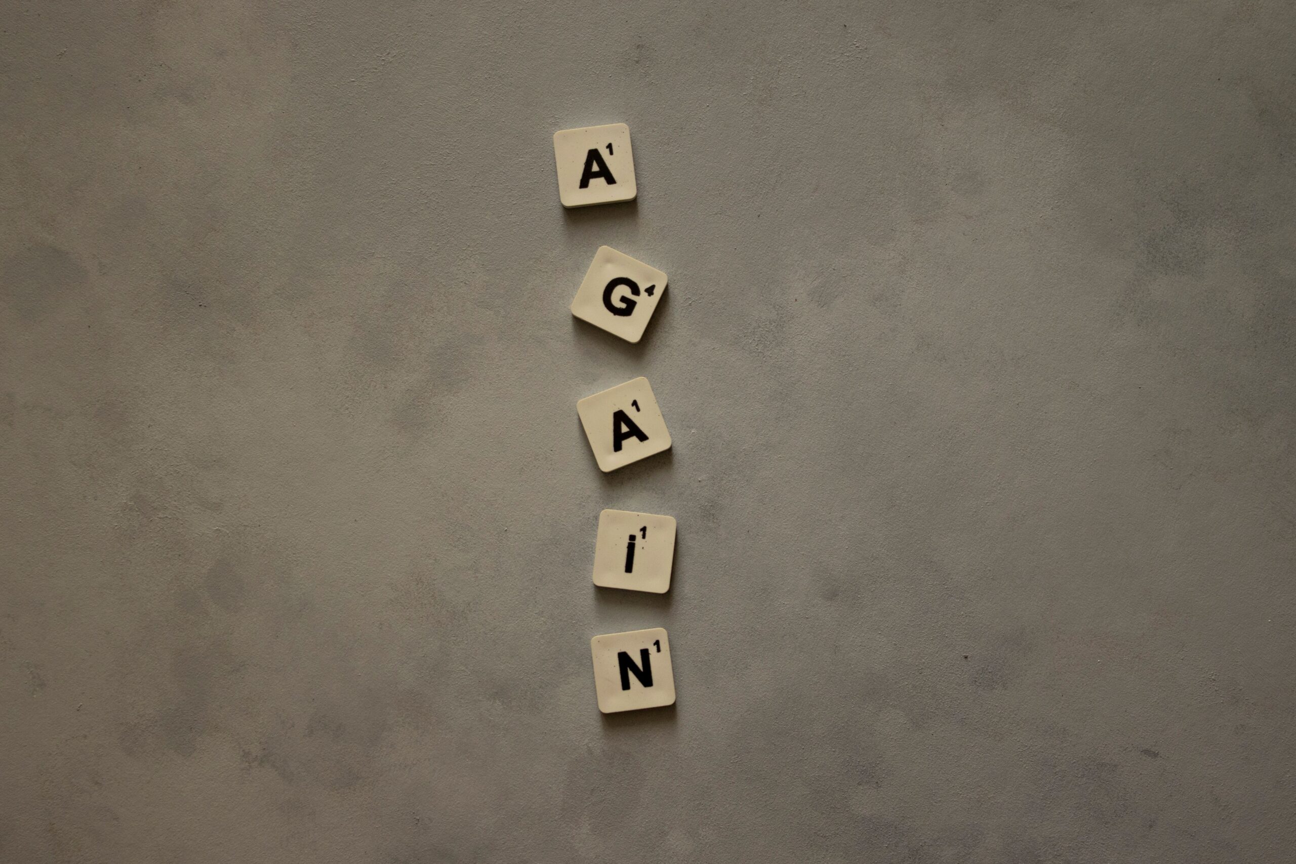 Scrabble letter tiles spelling 'Again' on textured concrete surface.