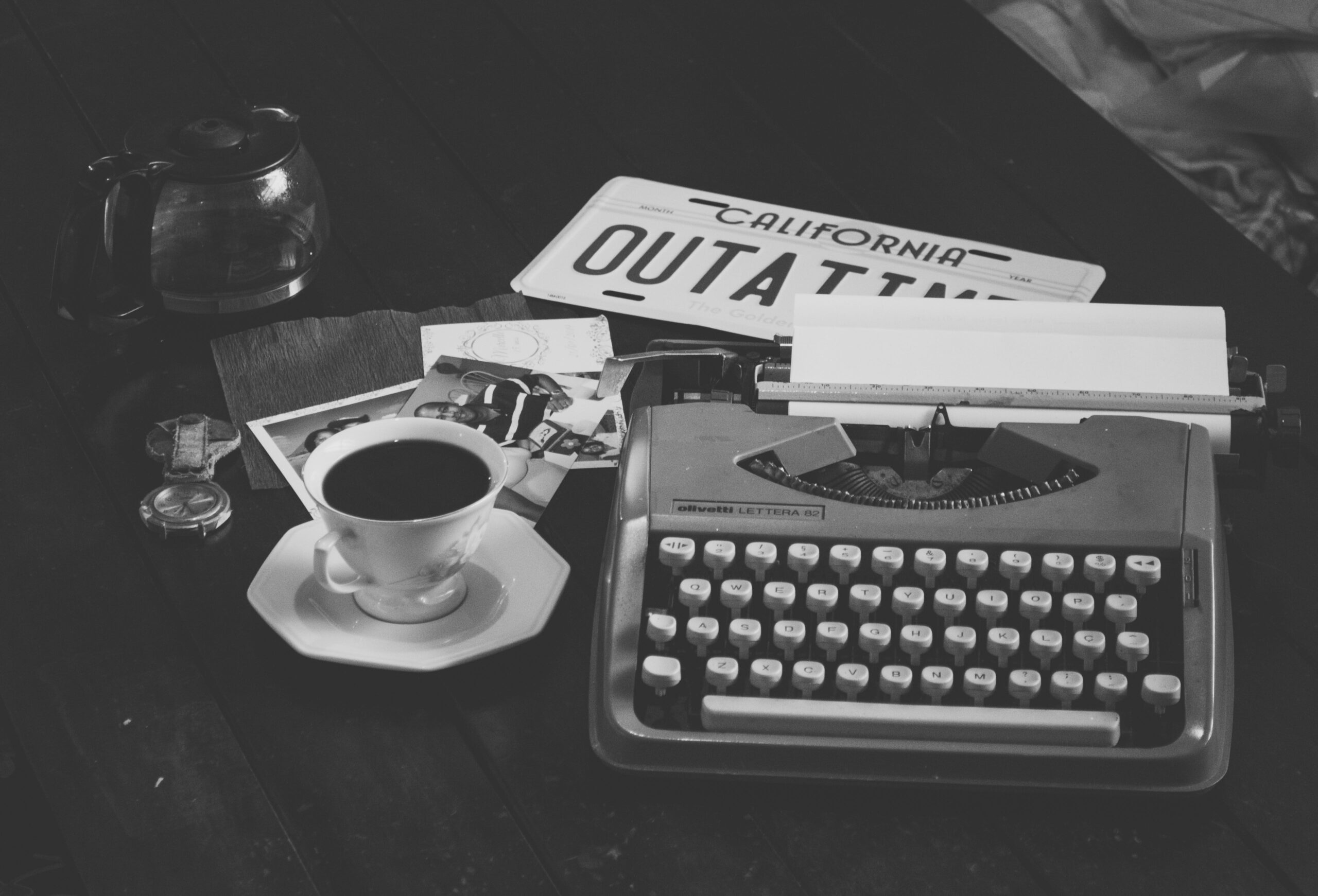 Black and white vintage setup featuring a typewriter with coffee, photos, and a California license plate.