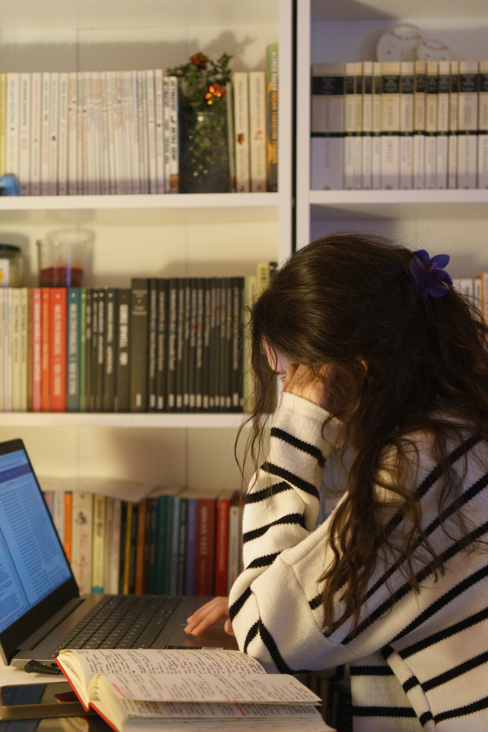 A woman in a striped sweater studies at home with a laptop and open notebook, surrounded by a bookshelf.