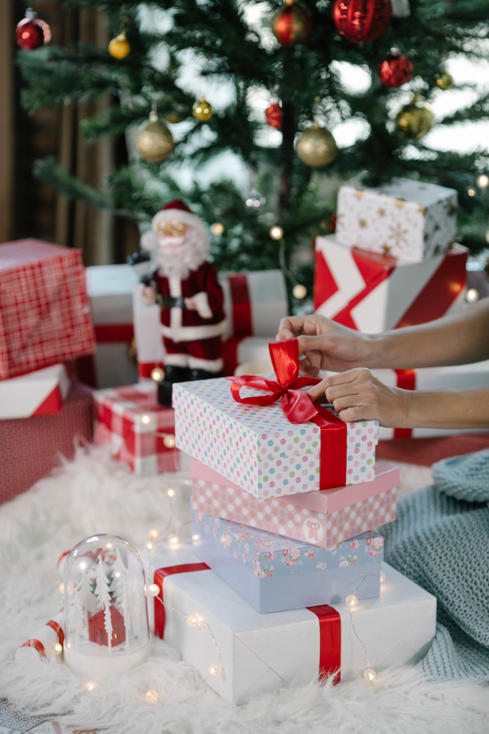 Hands wrapping Christmas presents with ribbons under a festive tree indoors.