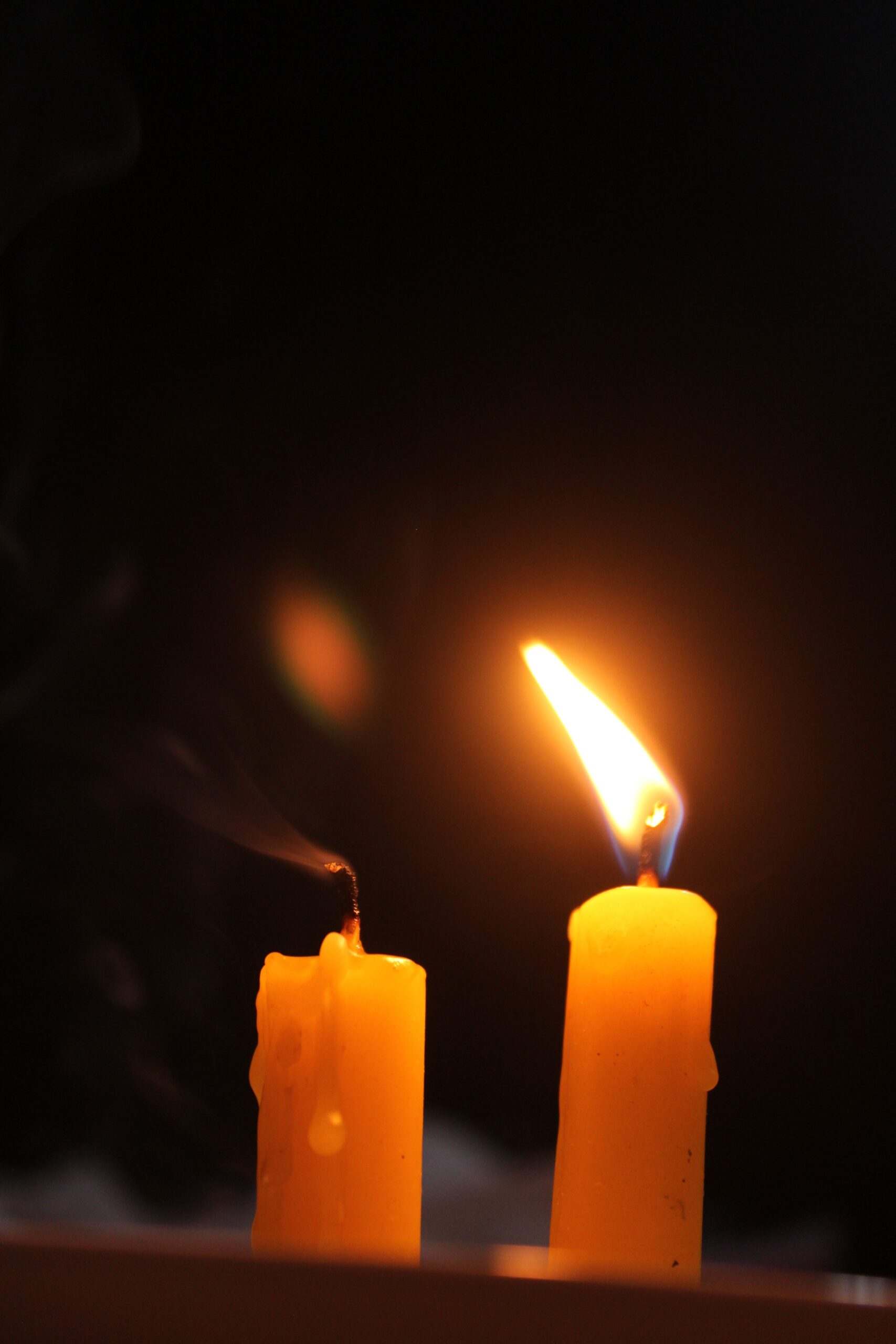 Two candles, one lit and one extinguished, against a dark backdrop, emitting a warm glow.