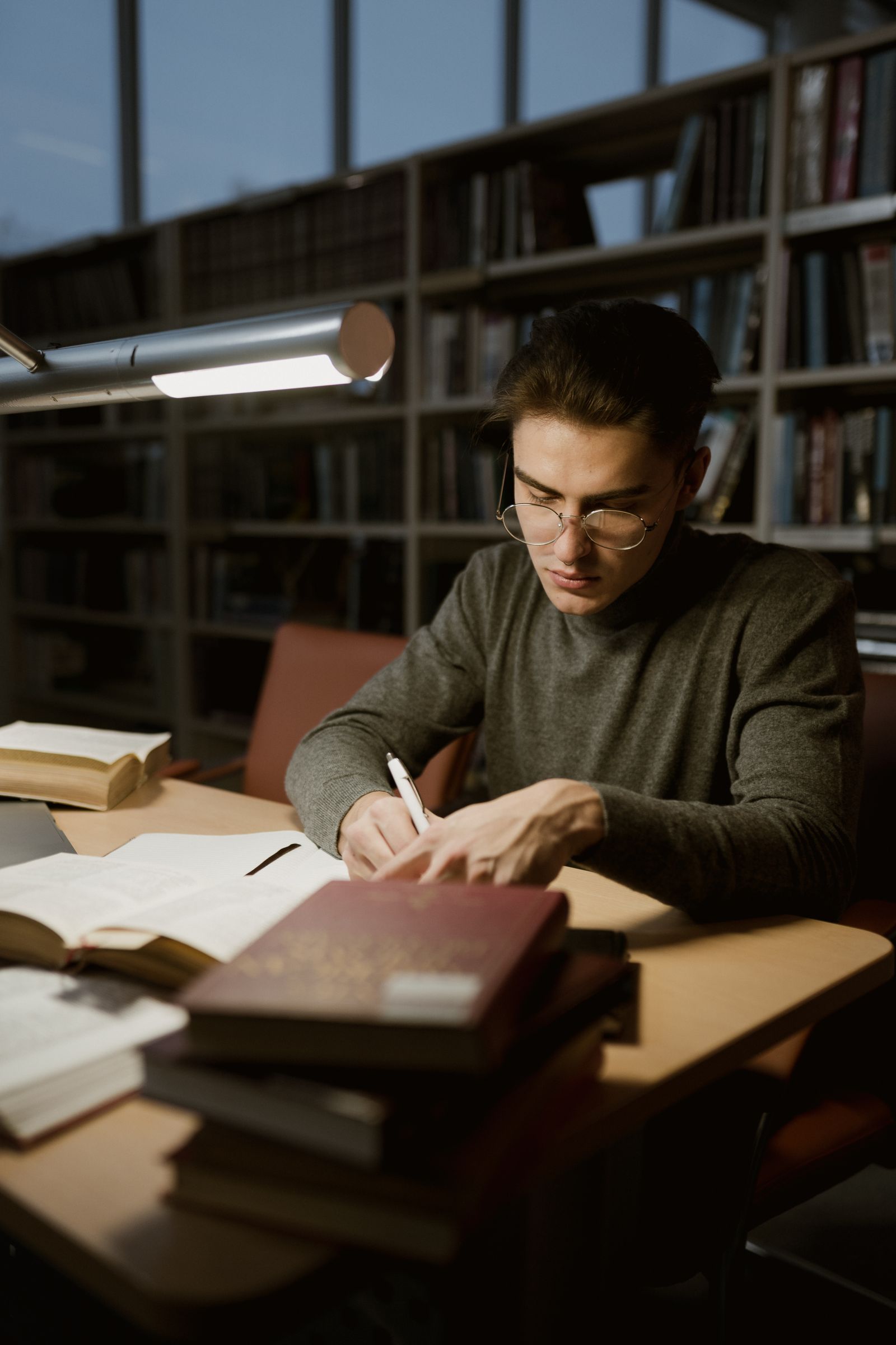 A young man in a library studying with books and taking notes under a desk lamp