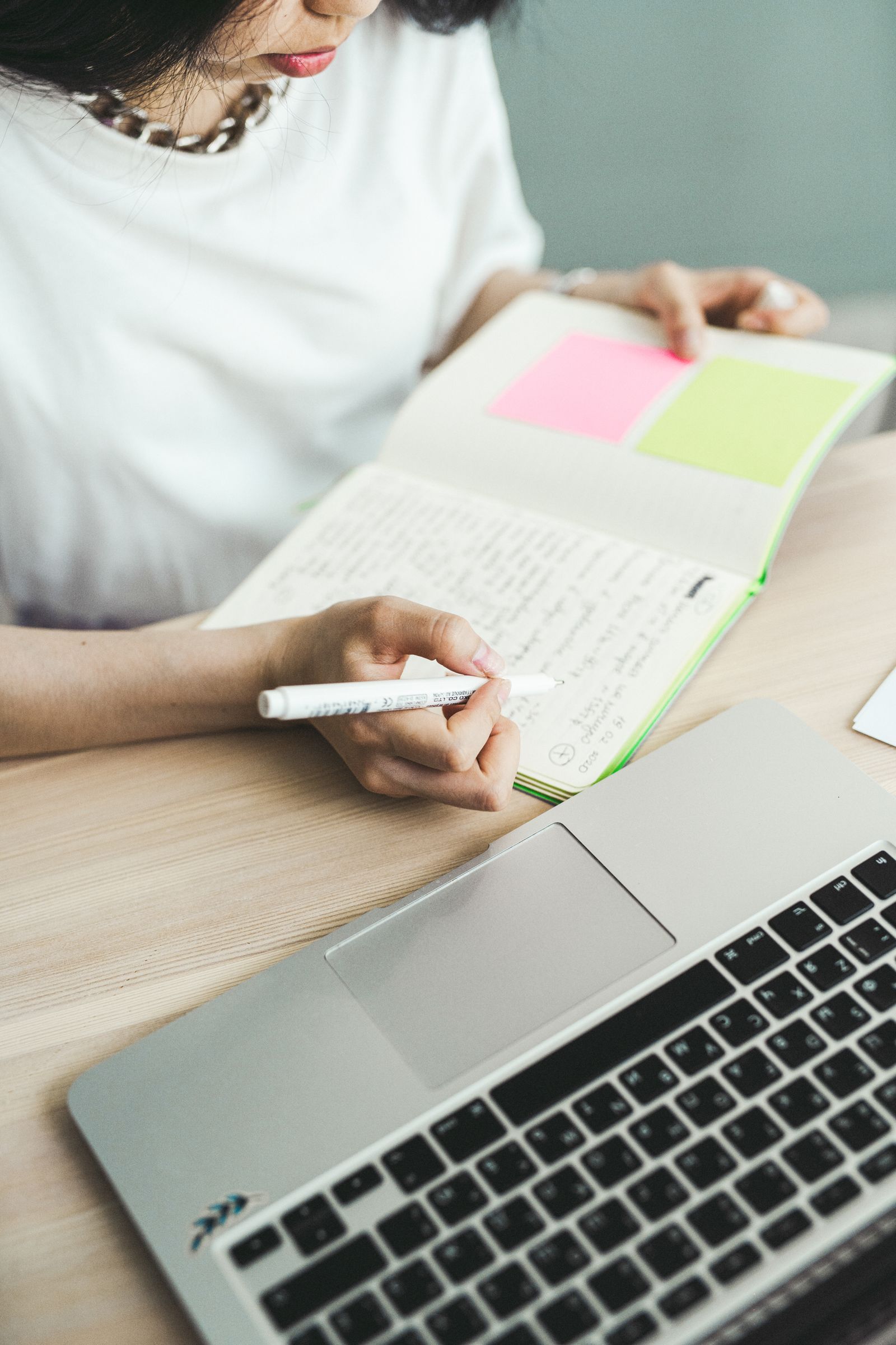 Close-up of a woman writing notes in a notebook beside a laptop on a wooden desk
