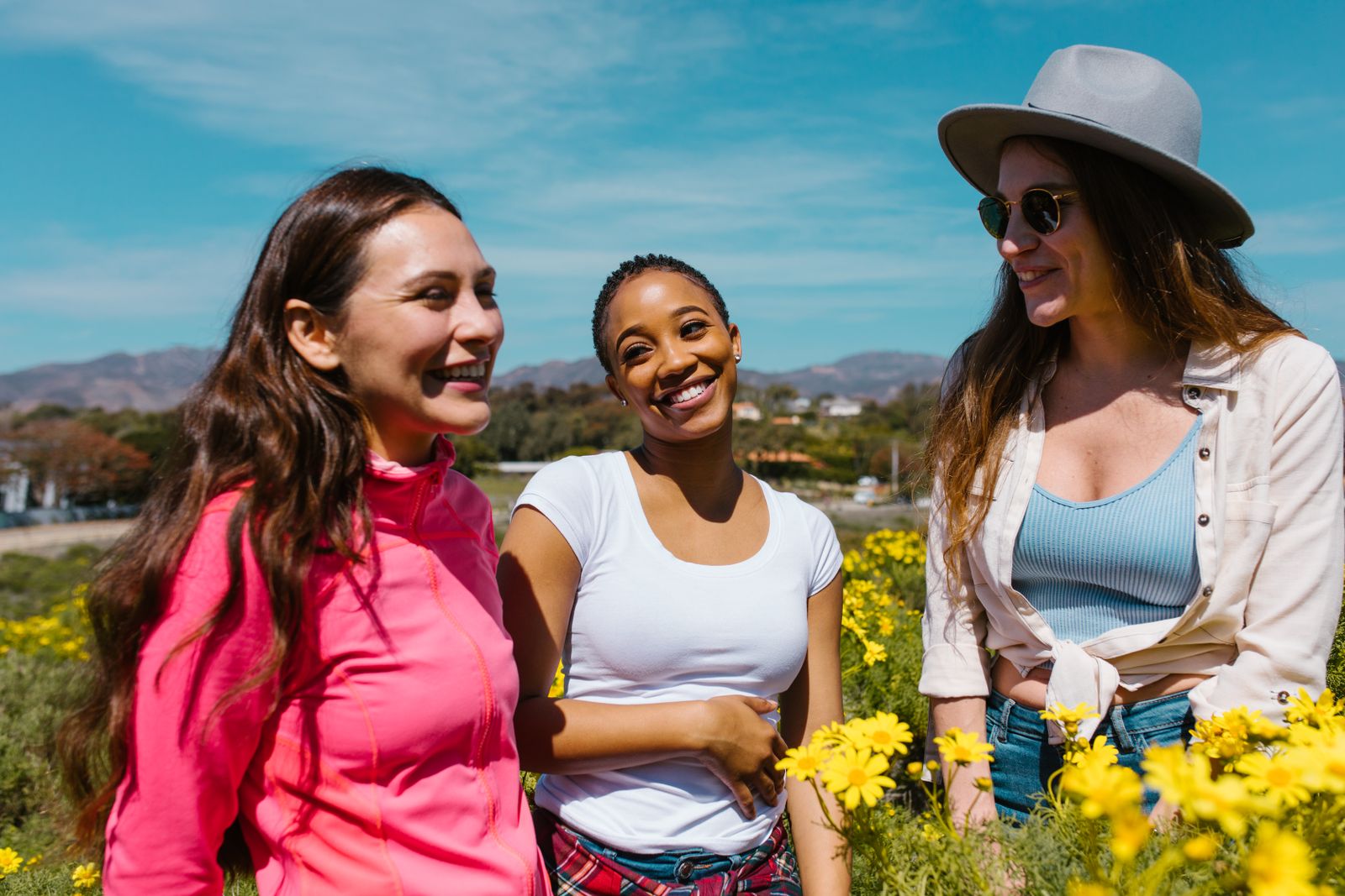 Three friends smiling and laughing together outdoors among yellow wildflowers on a sunny day