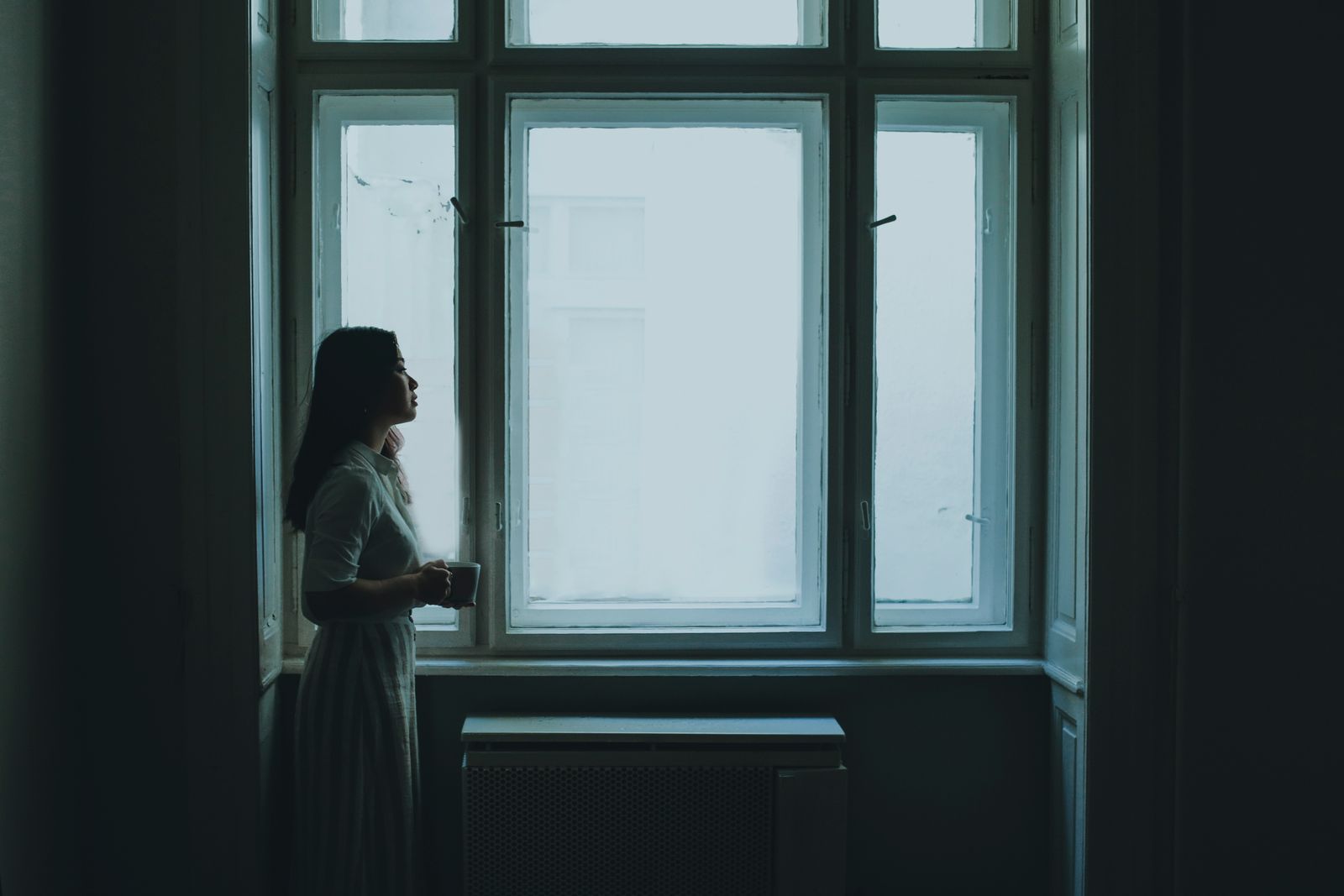 A young woman with long hair stands by a window holding a cup, creating a serene and contemplative mood