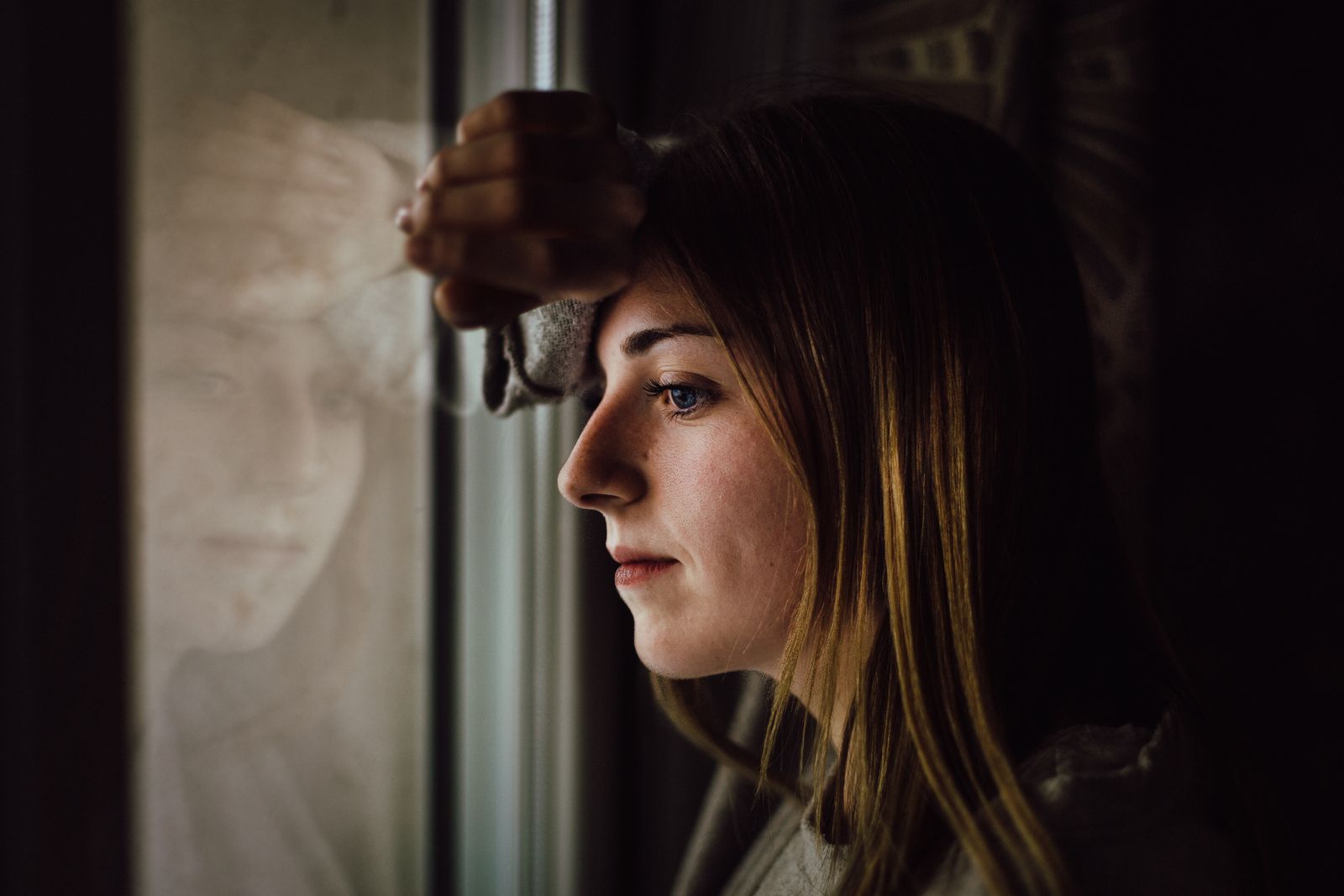 A contemplative young woman looks out a window with a reflective expression, indoors