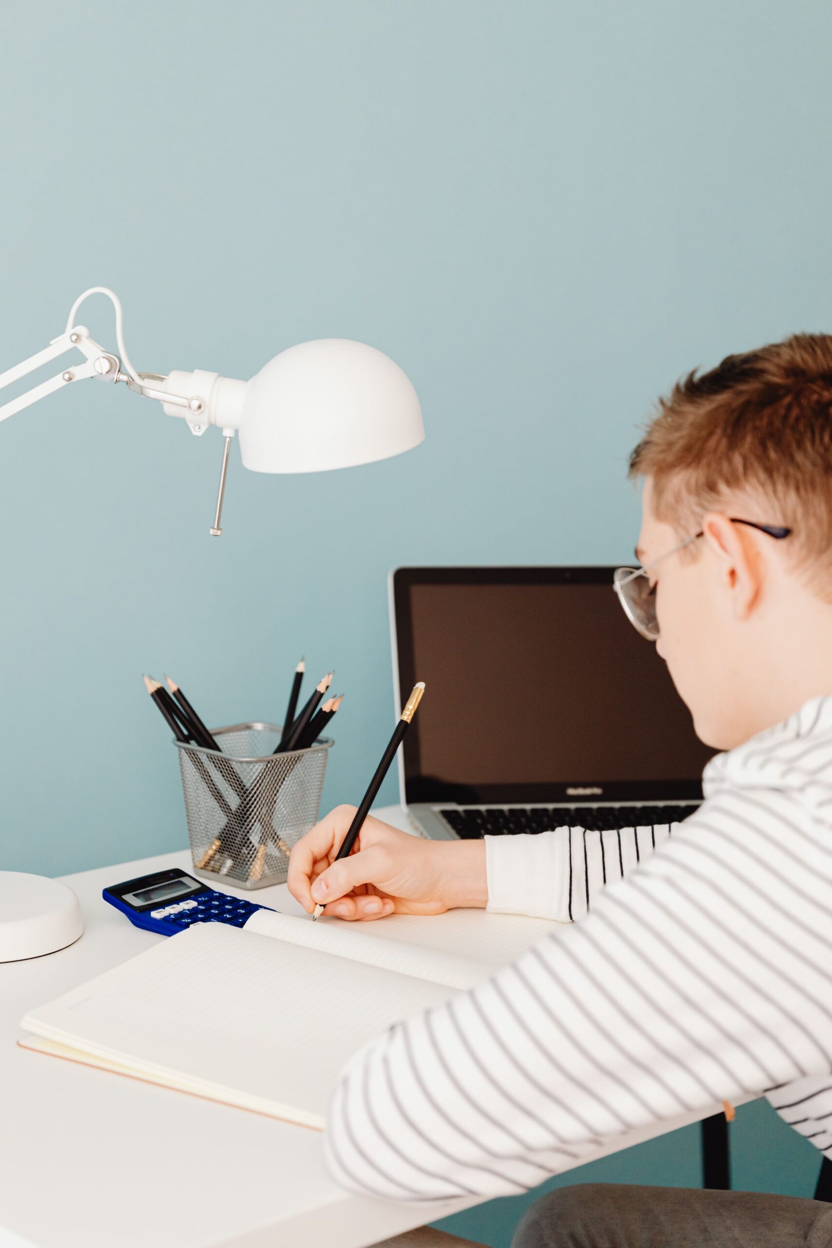 Man writing in a notebook at a desk with laptop and lamp