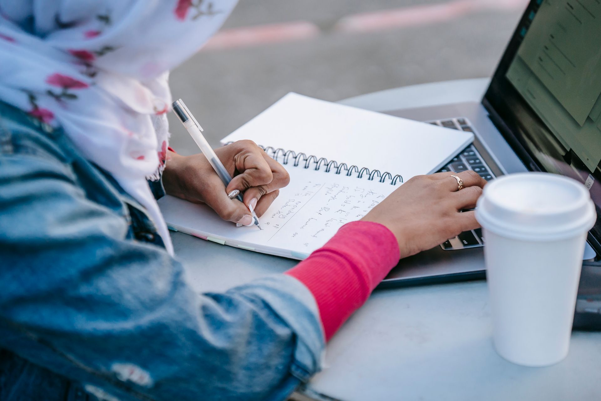 Muslim woman taking notes and using laptop outdoors, focused on research.