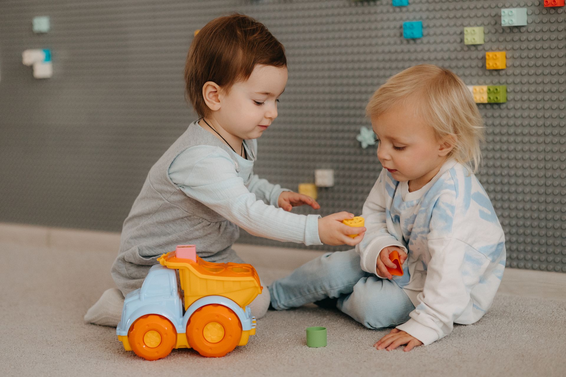 Two toddlers happily playing with a toy truck indoors, surrounded by colorful blocks.