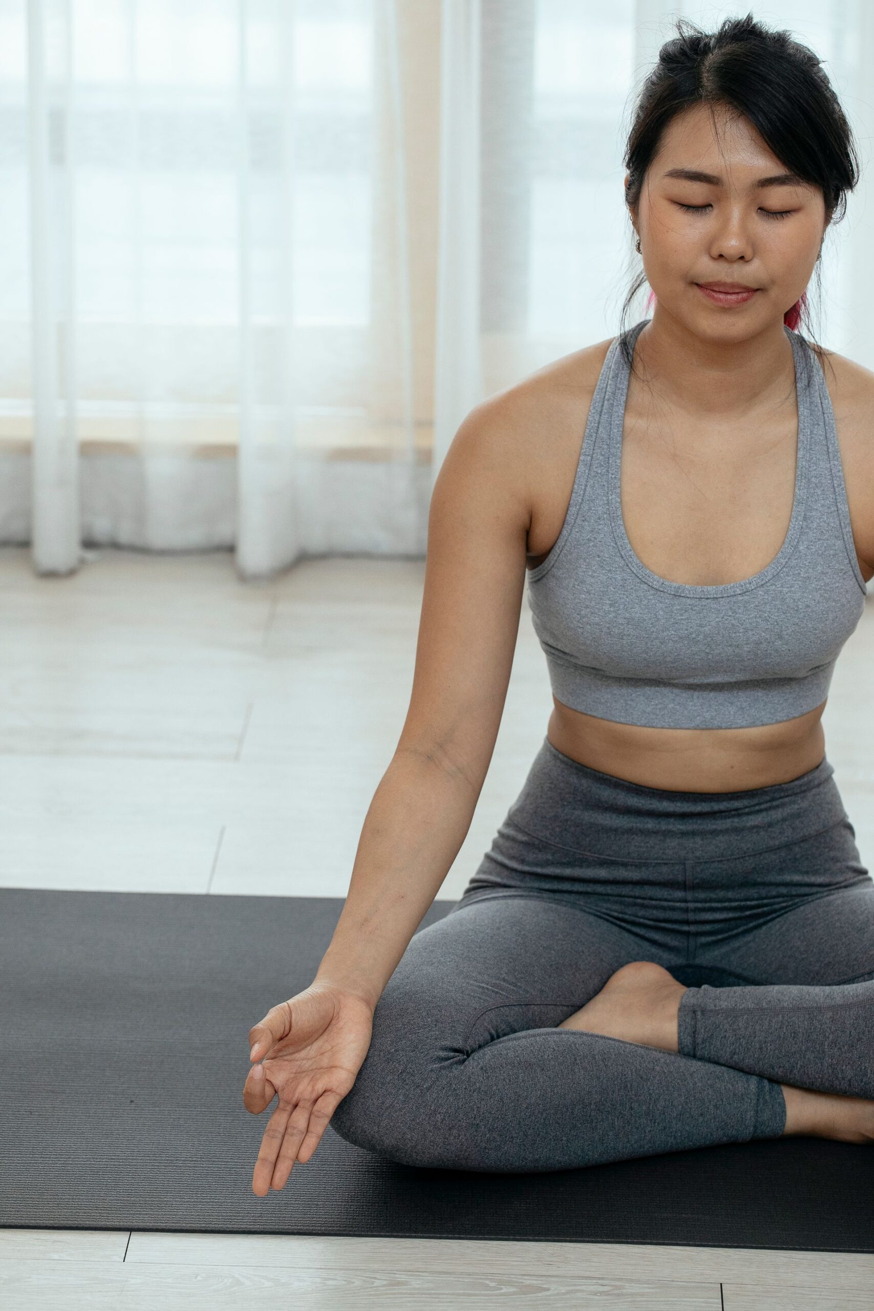 Asian woman in activewear sitting cross-legged, meditating on a yoga mat indoors.