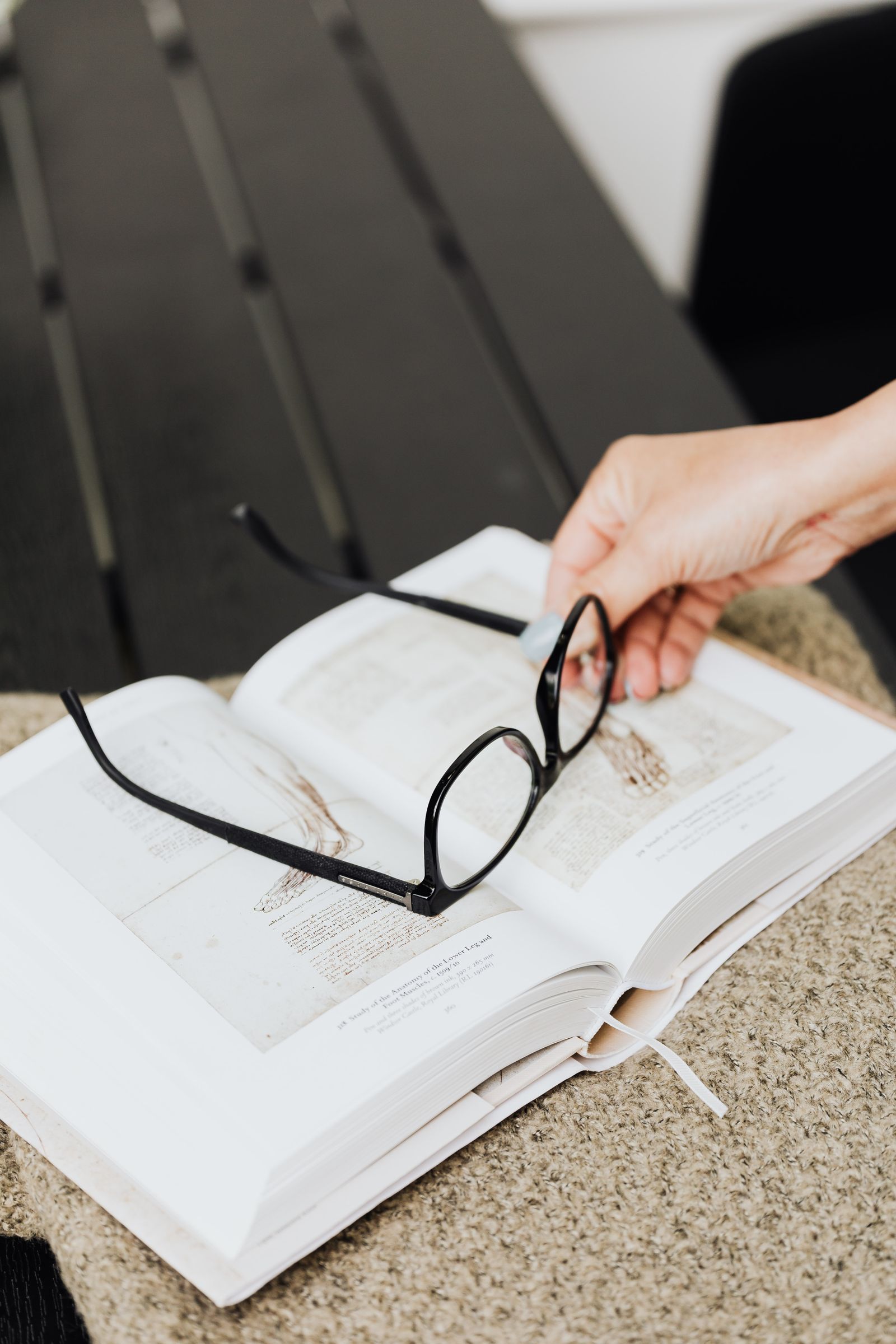A hand holds glasses over an open book on a table, capturing a moment of study