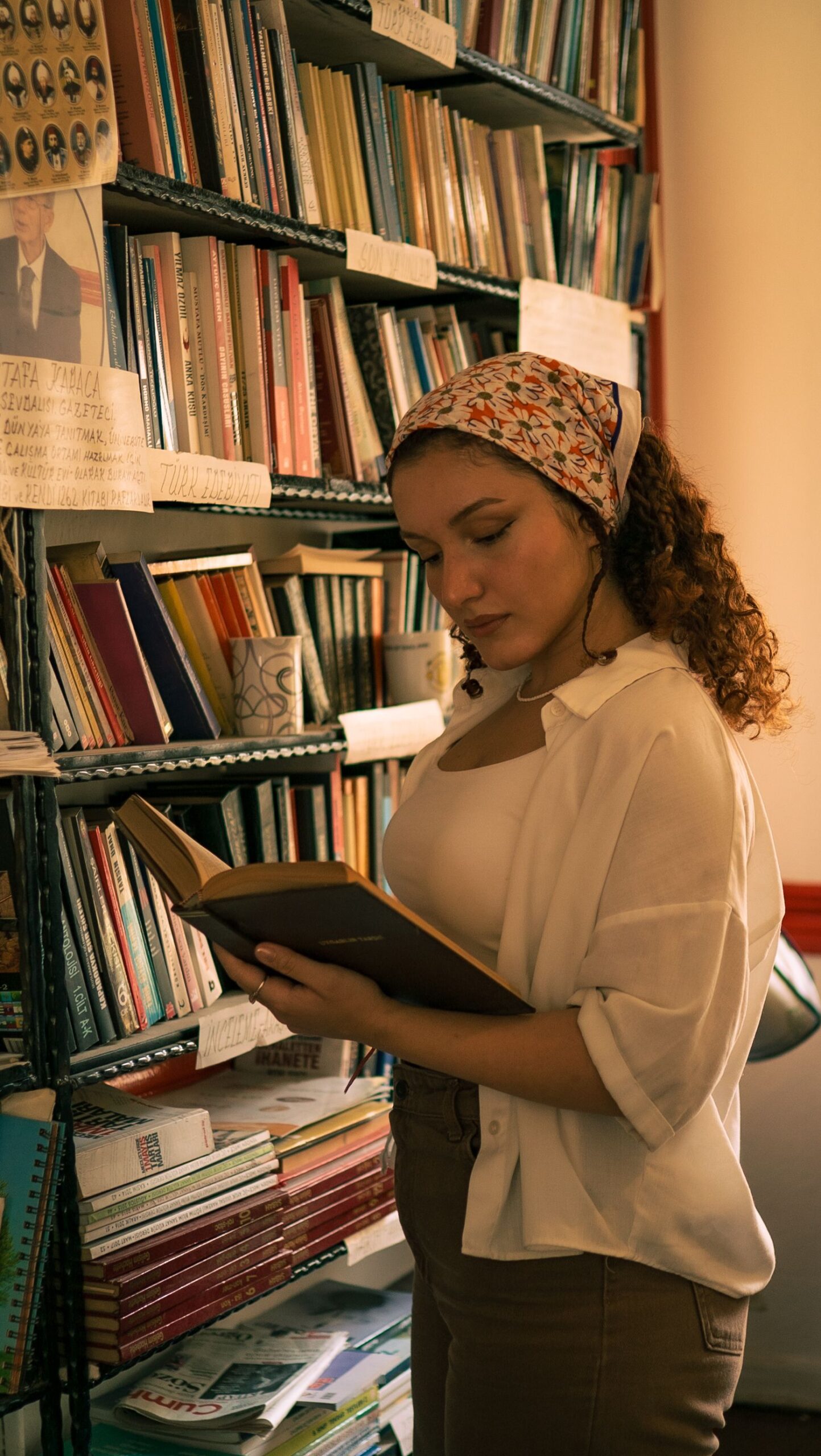 A thoughtful woman with a book stands among towering bookshelves in a dimly lit library