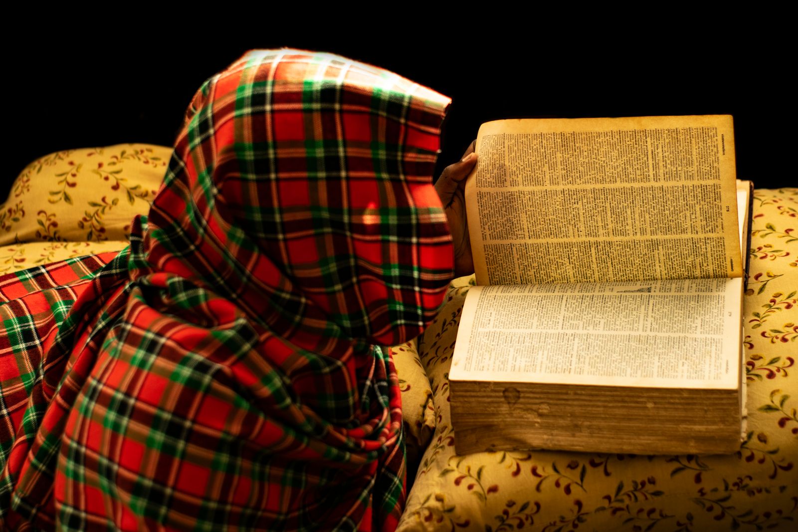 A person reading a book wrapped in a cozy plaid blanket, indoors, warm lighting