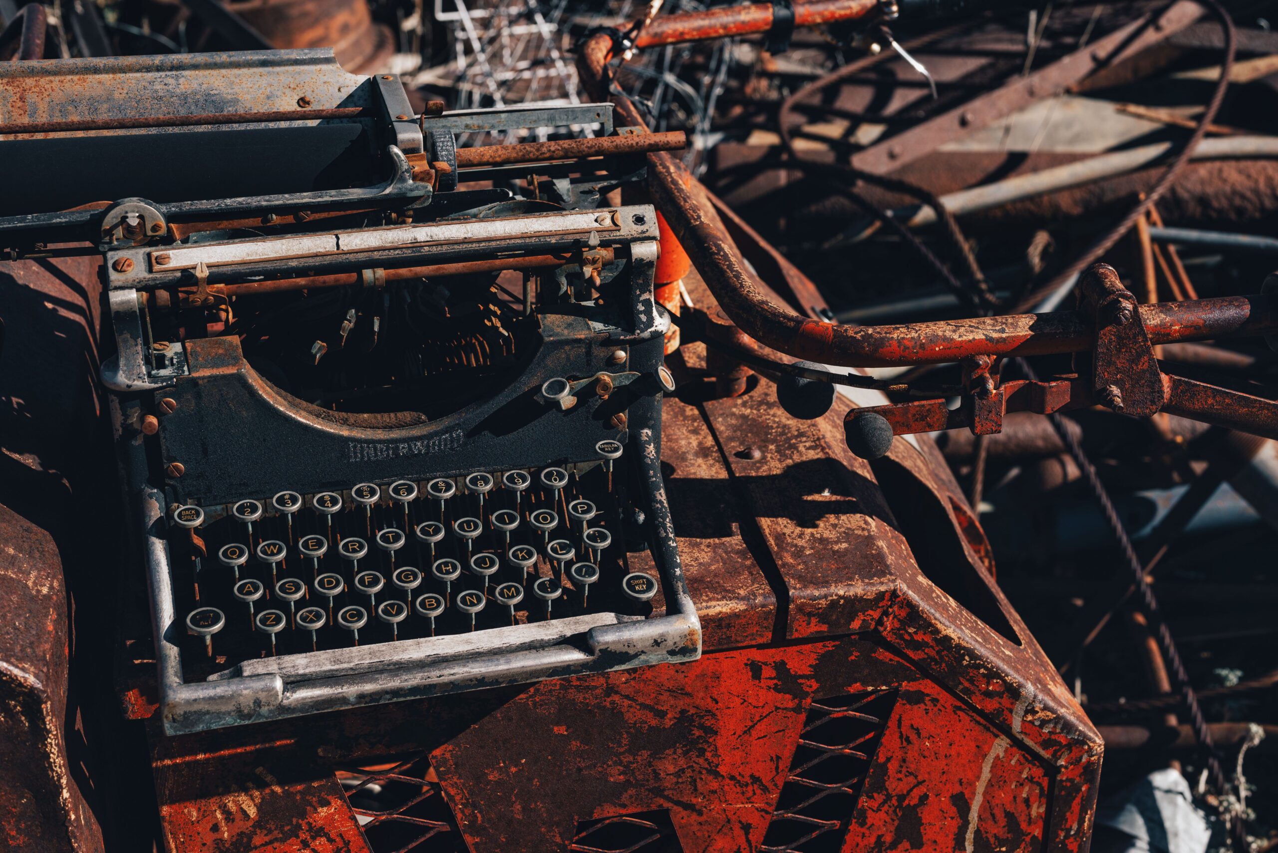 Rusty typewriter amidst metal scrap in a junkyard, bathed in harsh sunlight.