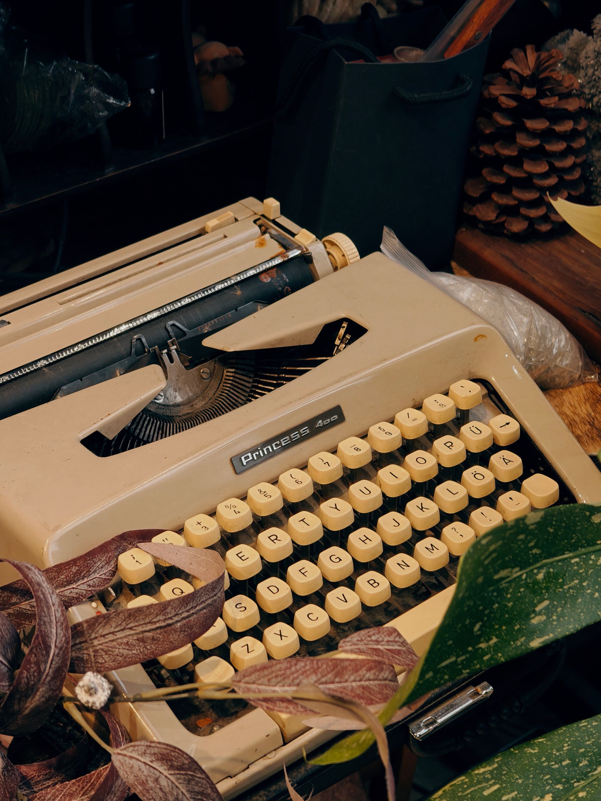 A classic beige typewriter sits amidst leaves and pine cones, exuding a vintage charm.