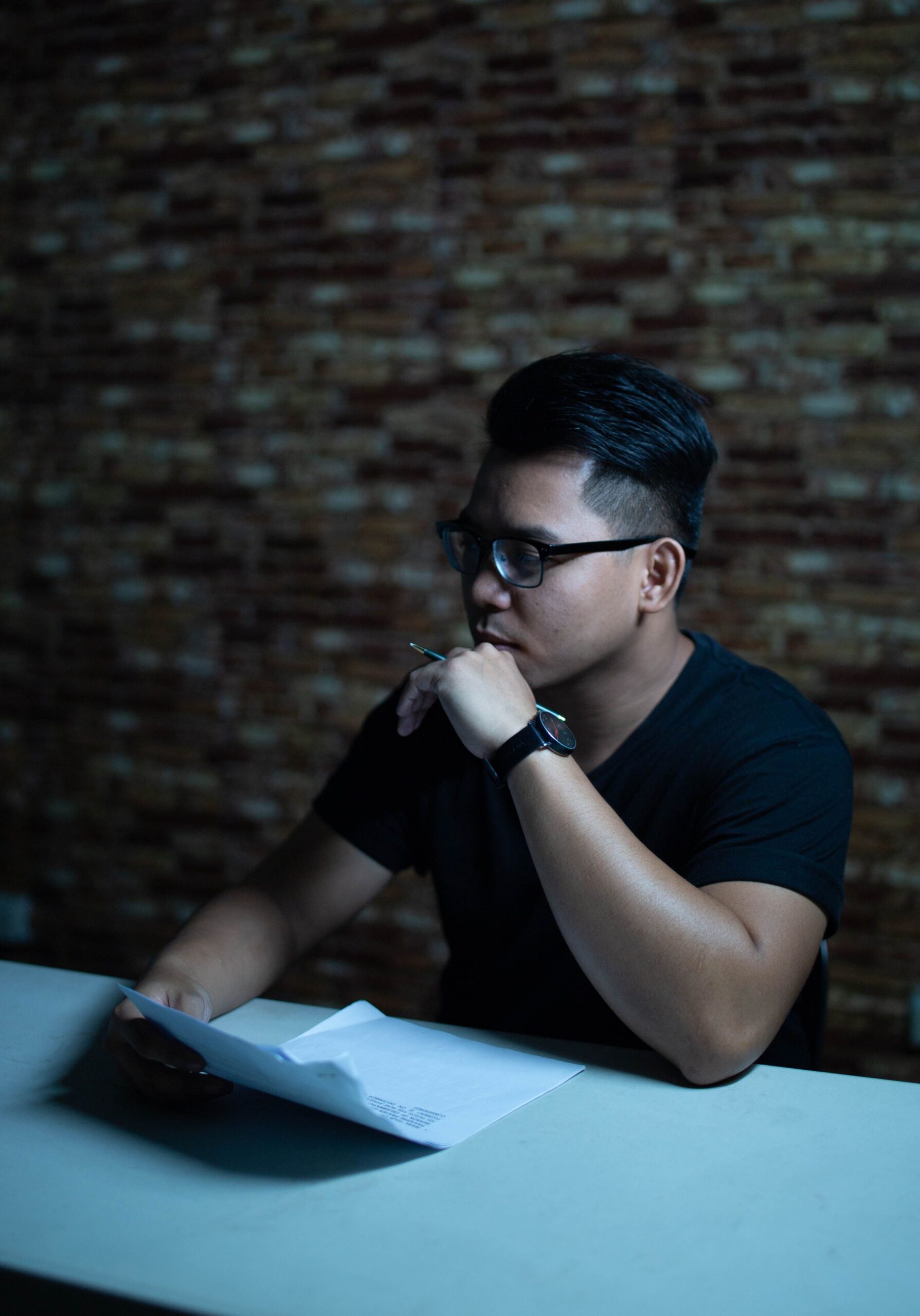 Man wearing glasses thinking with a pen at a table indoors